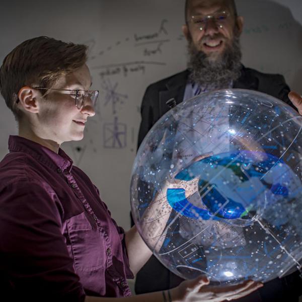 A professor stands behind a clear model of the milky way. Light shines up from beneath the model, onto the face of a student to whom he is explaining it.