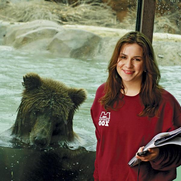 A student standing in front of a window. Behind them a grizzly bear is eating a fish.